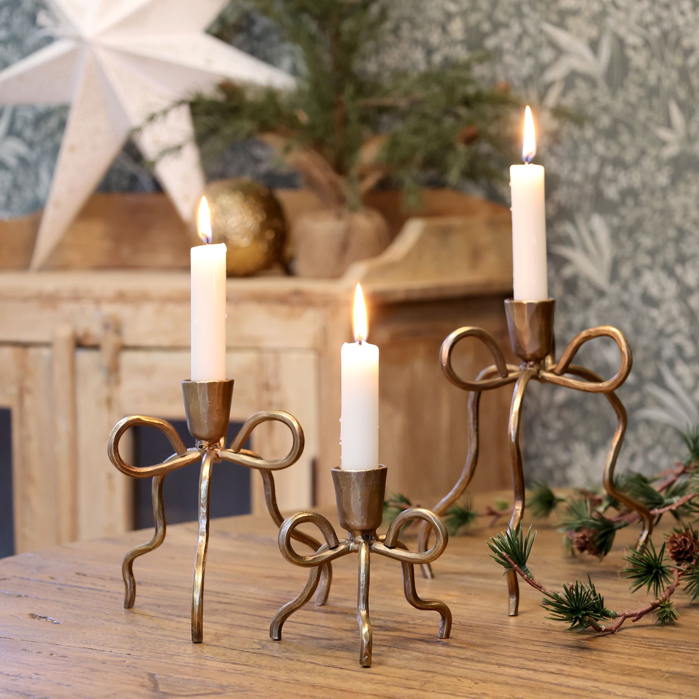 Gold bow candlestick holders with white taper candles, available in three sizes, styled on a wooden table with festive greenery