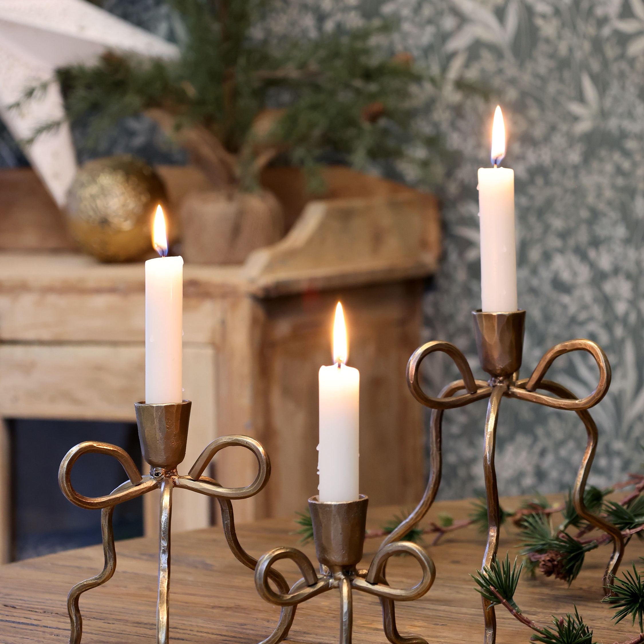 Gold bow candlestick holders with white taper candles, available in three sizes, styled on a wooden table with festive greenery