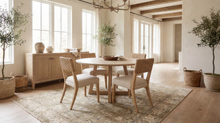 A round wooden dining table with four woven-back chairs in a bright, modern dining room with large windows, a sideboard, and potted plants.