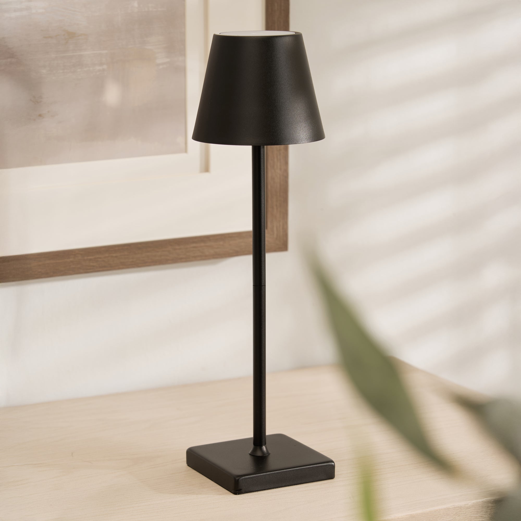 A sleek black wireless lamp with a conical shade placed on a square base, situated on a wooden surface with a wall and window in the background.