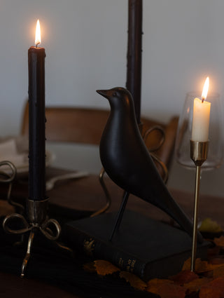 Black bird figurine between two lit candles on a reflective surface.
