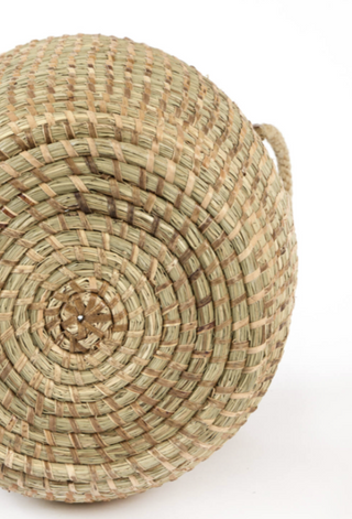 A close-up of a round woven basket made from natural straw or seagrass, showing the detailed spiral weave pattern and a small loop handle on the right side, set against a plain white background.