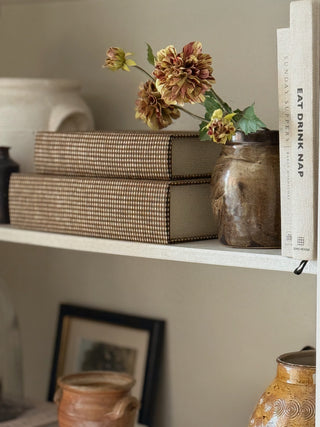 A styled interior shelf scene with two woven-bound books, a rustic ceramic jar holding dried flowers, a white book titled "EAT DRINK NAP," and other home decor objects against a warm beige wall.
