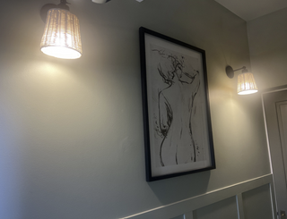 A framed black and white line art print of a female figure hangs on a light gray wall between two warm-toned wall sconces.