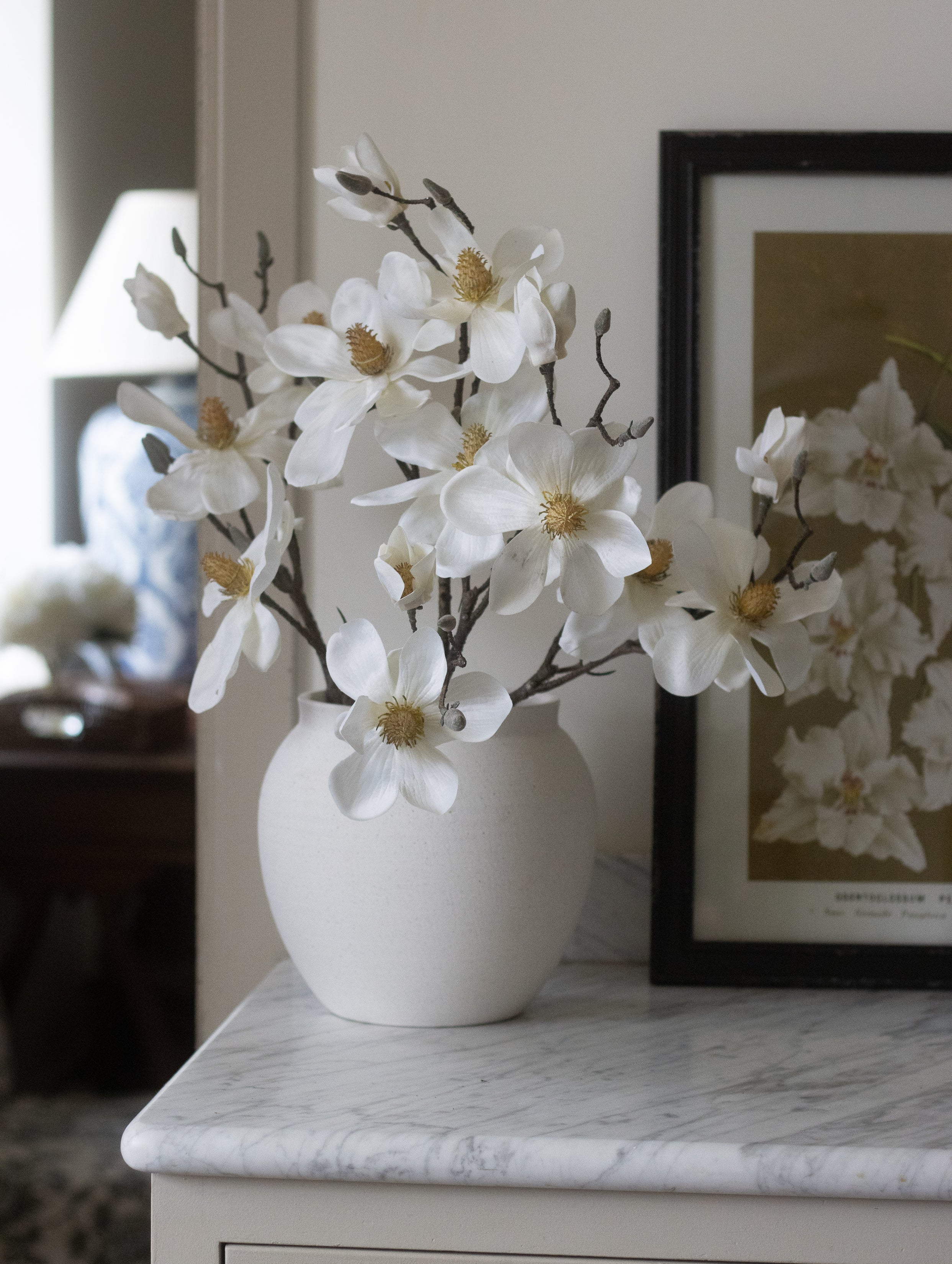 White vase with artificial flowers on a marble surface next to a framed picture