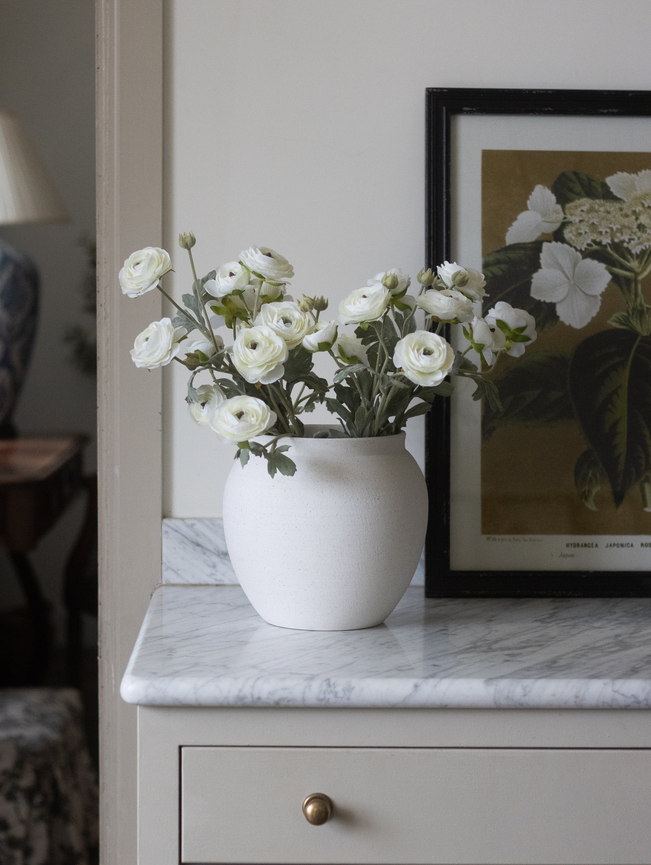 White flowers in a vase on a marble surface with a framed picture in the background