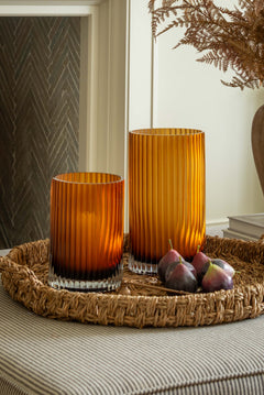 Two orange glass candle holders on a woven tray with figs, against a neutral wall.