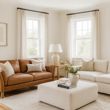 A bright, modern living room with white walls and large windows letting in natural light. At the centre is a vintage brown leather two-seater sofa with neutral throw pillows, paired with a cream fabric ottoman coffee table styled with books and a ceramic vase of greenery. A light fabric sofa sits opposite, completing the cosy, timeless Studio McGee inspired aesthetic with layered textures, warm wood flooring, and minimal, elegant décor.