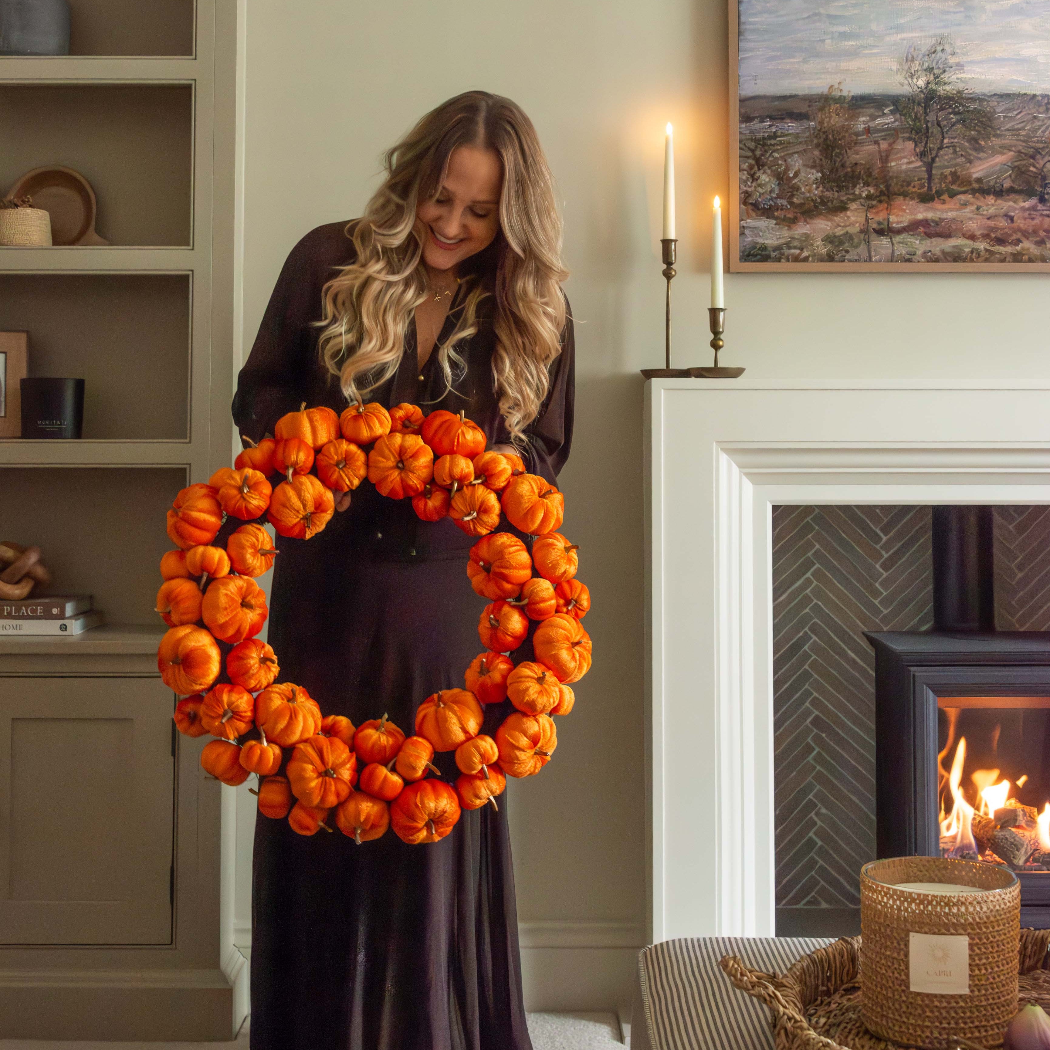 Woman holding a large orange pumpkin wreath in a cozy living room.