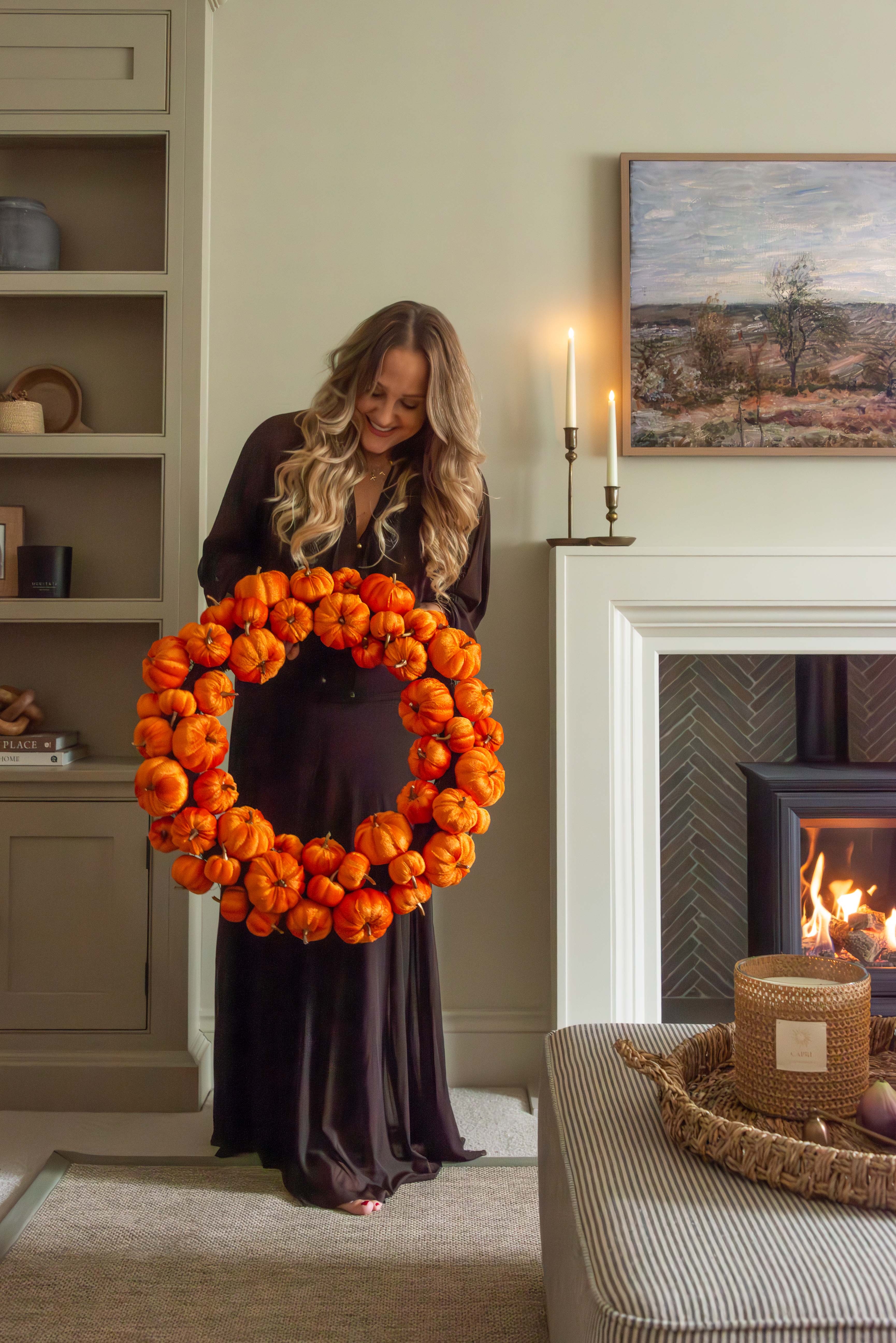 Woman holding a large orange pumpkin wreath in a cozy living room.