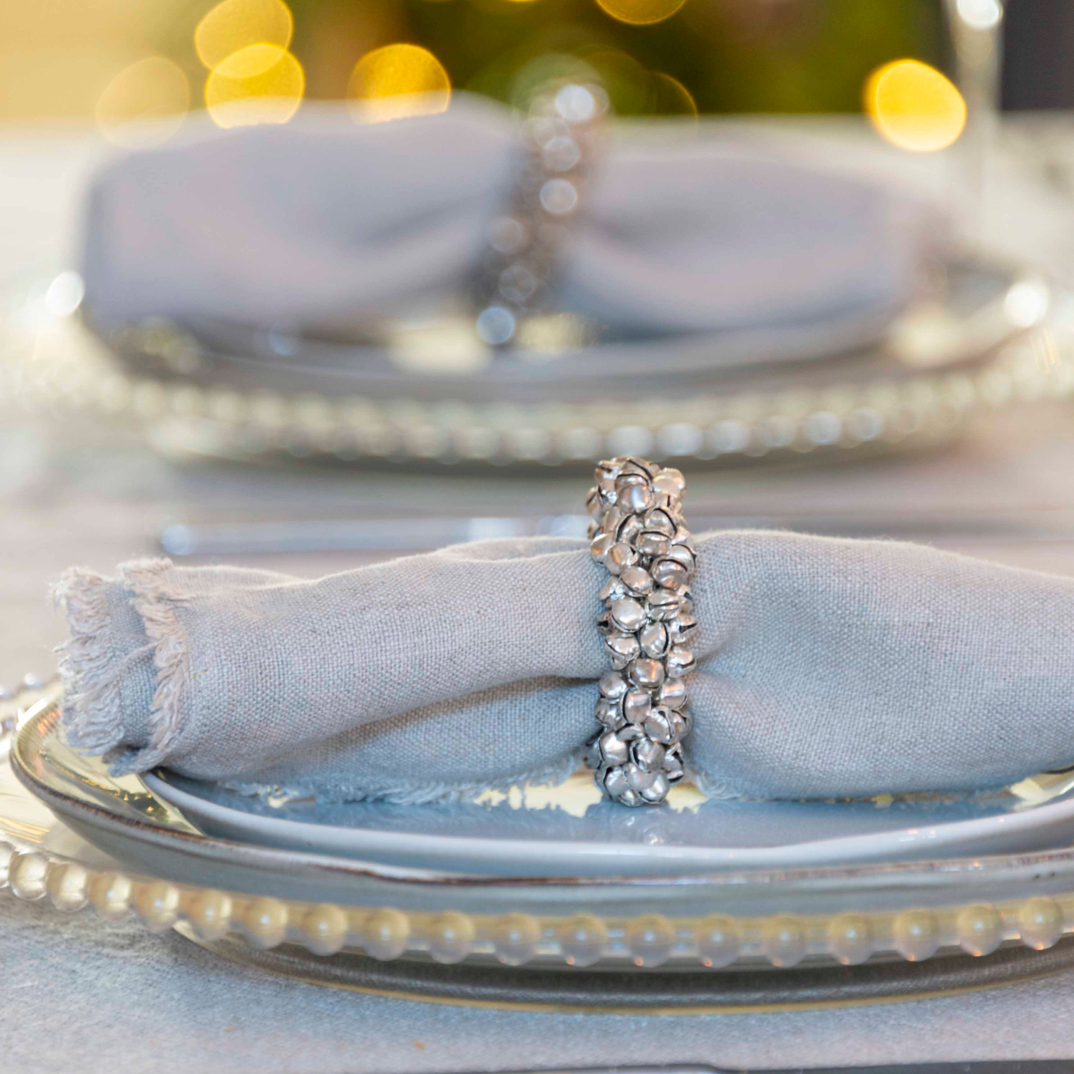A silver napkin ring with a bell shape displayed on a napkin, part of a table setting with dishes and cutlery in the background.