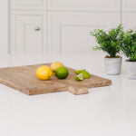 A large rectangular breadboard made of recycled elm. Shown with some whole lemons and wedges of lime resting near the protruding handle