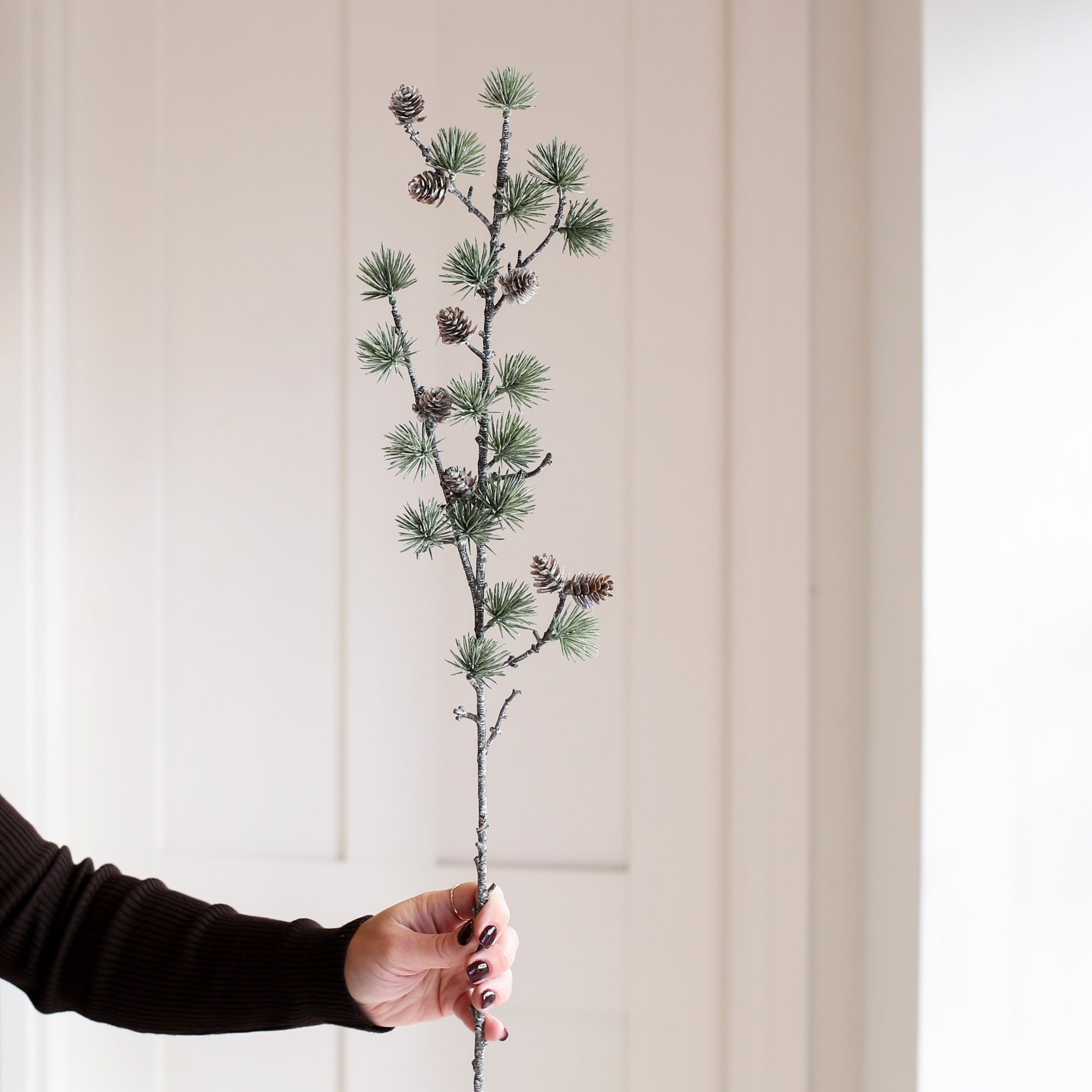 Hand holding a Charles Ted Frosted Cedar Spray with lifelike greenery and pine cones against a neutral background