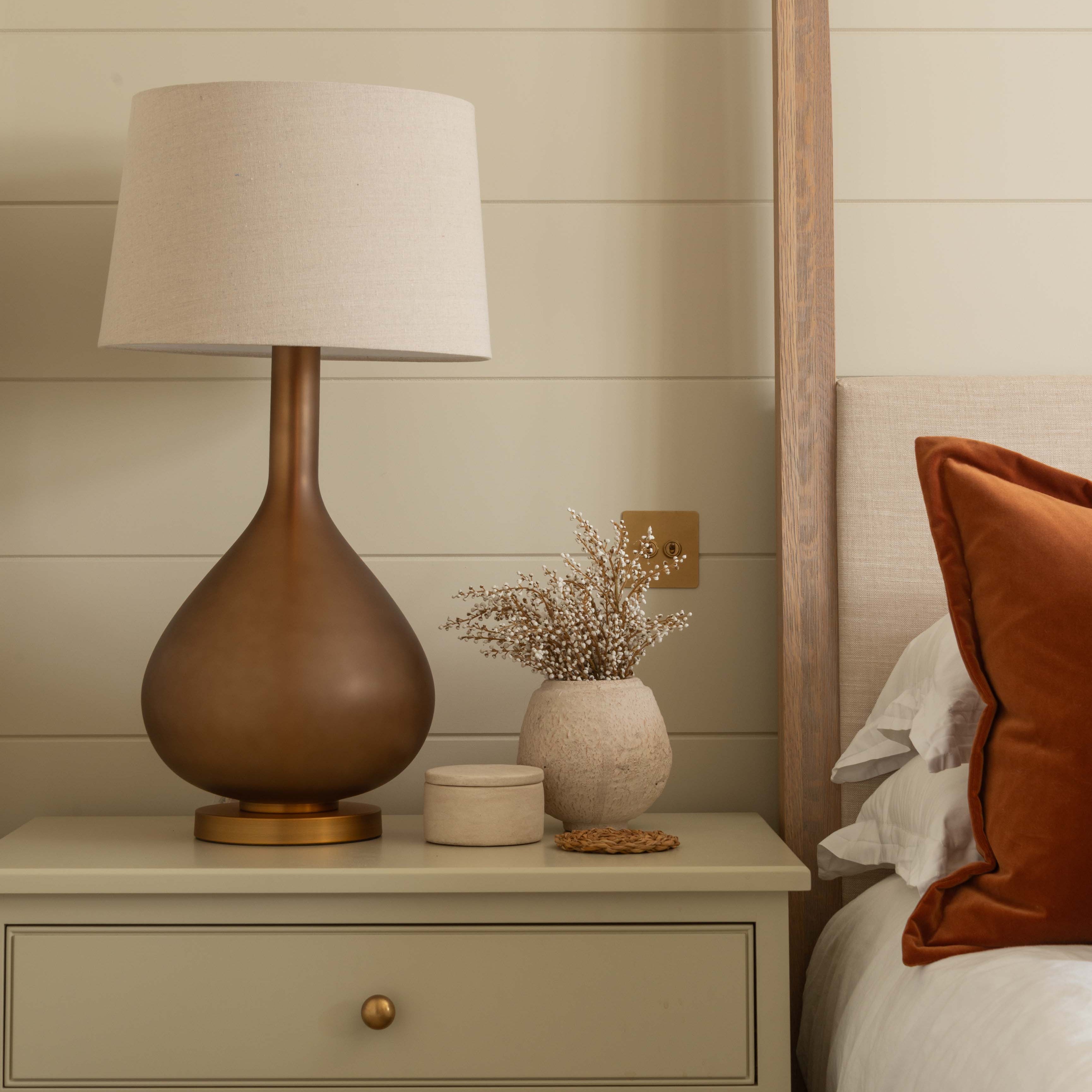 Nighstand with lamp and decorative items next to a bed with orange pillow.