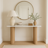 A fluted console table with beige leg detailing, accompanied by a lamp, a mirror above, and decorative items on the table surface, set against a neutral wall in a home environment.