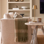 A beige wooden tripod side table in a living room setting, with a lamp and a book on top of it, featuring a rustic finish.