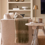 A beige wooden tripod side table in a living room setting, with a lamp and a book on top of it, featuring a rustic finish.