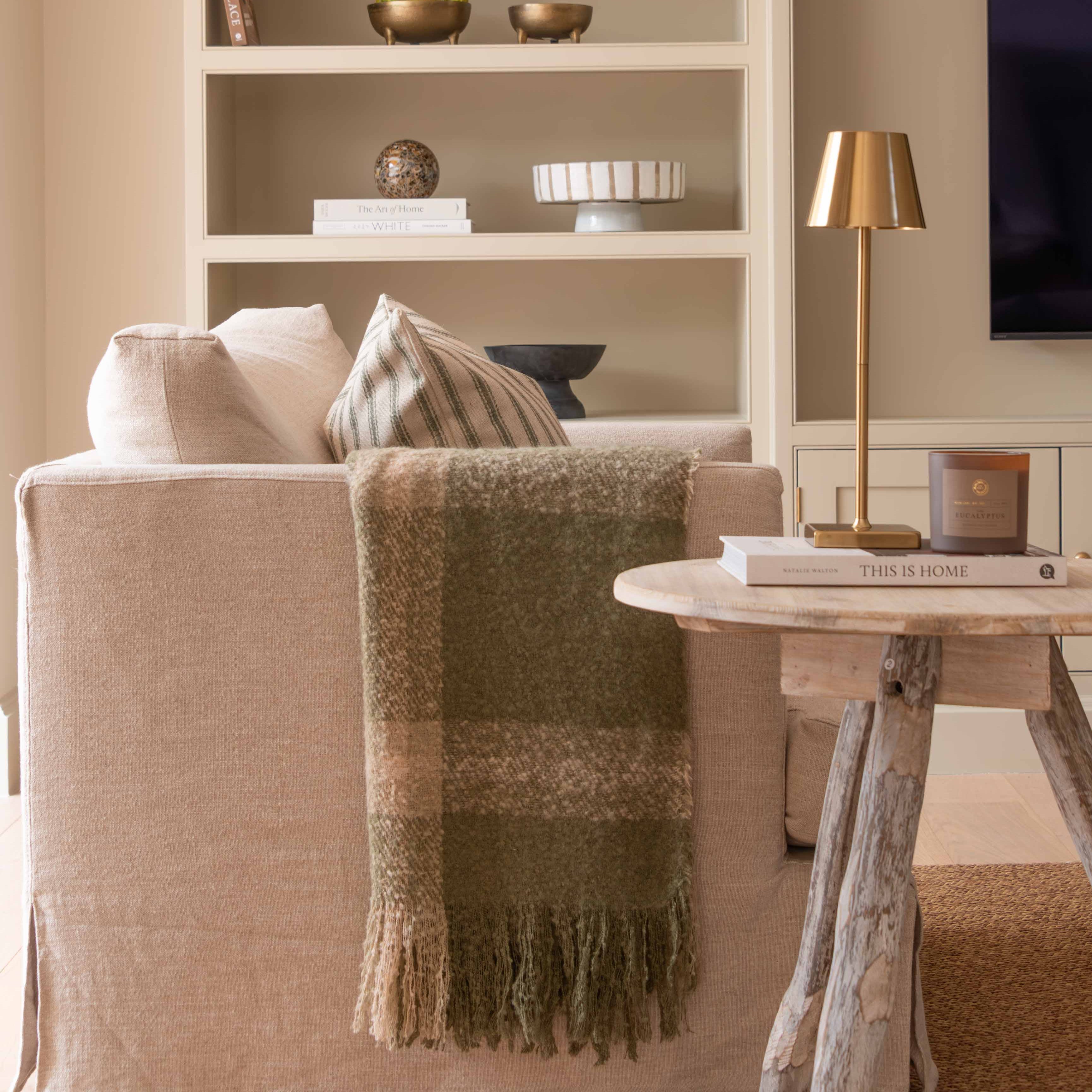 A beige wooden tripod side table in a living room setting, with a lamp and a book on top of it, featuring a rustic finish.