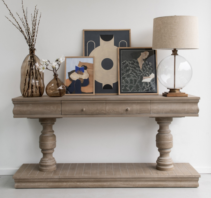 A gray effect console table with two drawers, placed against a white wall and styled with decorative items on top, including a lamp and framed art.