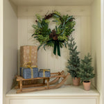 Decorative setup with a wreath, gift boxes, and small trees inside a white cabinet.
