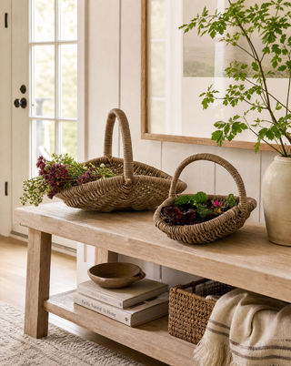 Two woven wicker baskets with handles, one larger and one smaller, styled on a light wood bench in a bright, neutral-toned room. The baskets are filled with green and purple flowers, with a potted plant and books in the background.