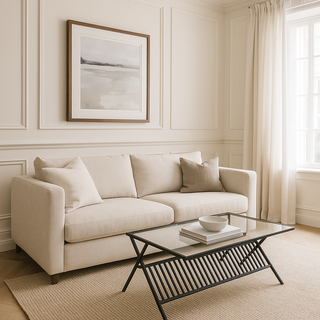 A sleek glass coffee table with a black iron frame, positioned in a well-lit room with a beige sofa, a framed picture on the wall, and a light-colored carpet.
