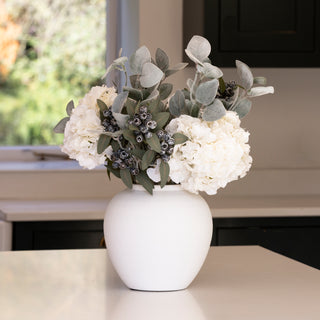 Frosted faux eucalyptus stems arranged with white hydrangeas in a round white vase