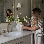 Woman arranging faux real touch white ranunculus bouquet in a white vase on a bathroom vanity
