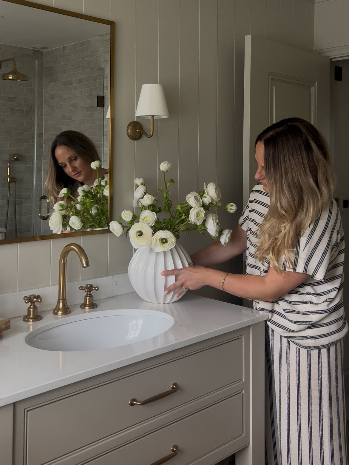 Woman arranging faux real touch white ranunculus bouquet in a white vase on a bathroom vanity