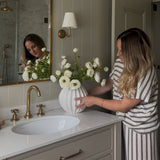 Woman arranging faux real touch white ranunculus bouquet in a white vase on a bathroom vanity