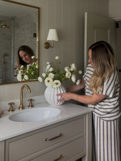 Woman arranging faux real touch white ranunculus bouquet in a white vase on a bathroom vanity