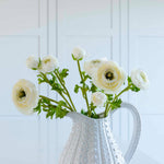 White ceramic pitcher holding realistic faux white ranunculus flowers with green leaves