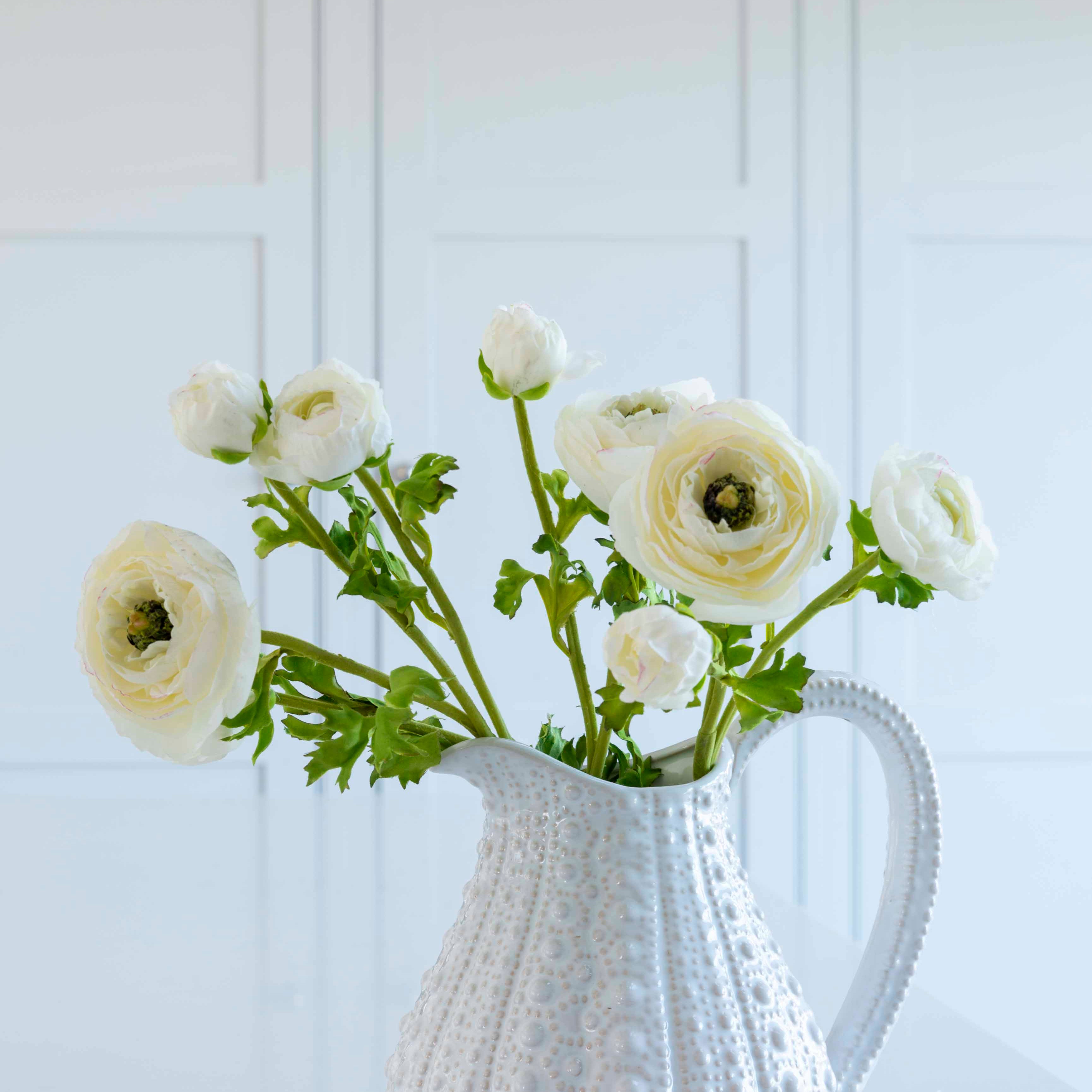 White ceramic pitcher holding realistic faux white ranunculus flowers with green leaves