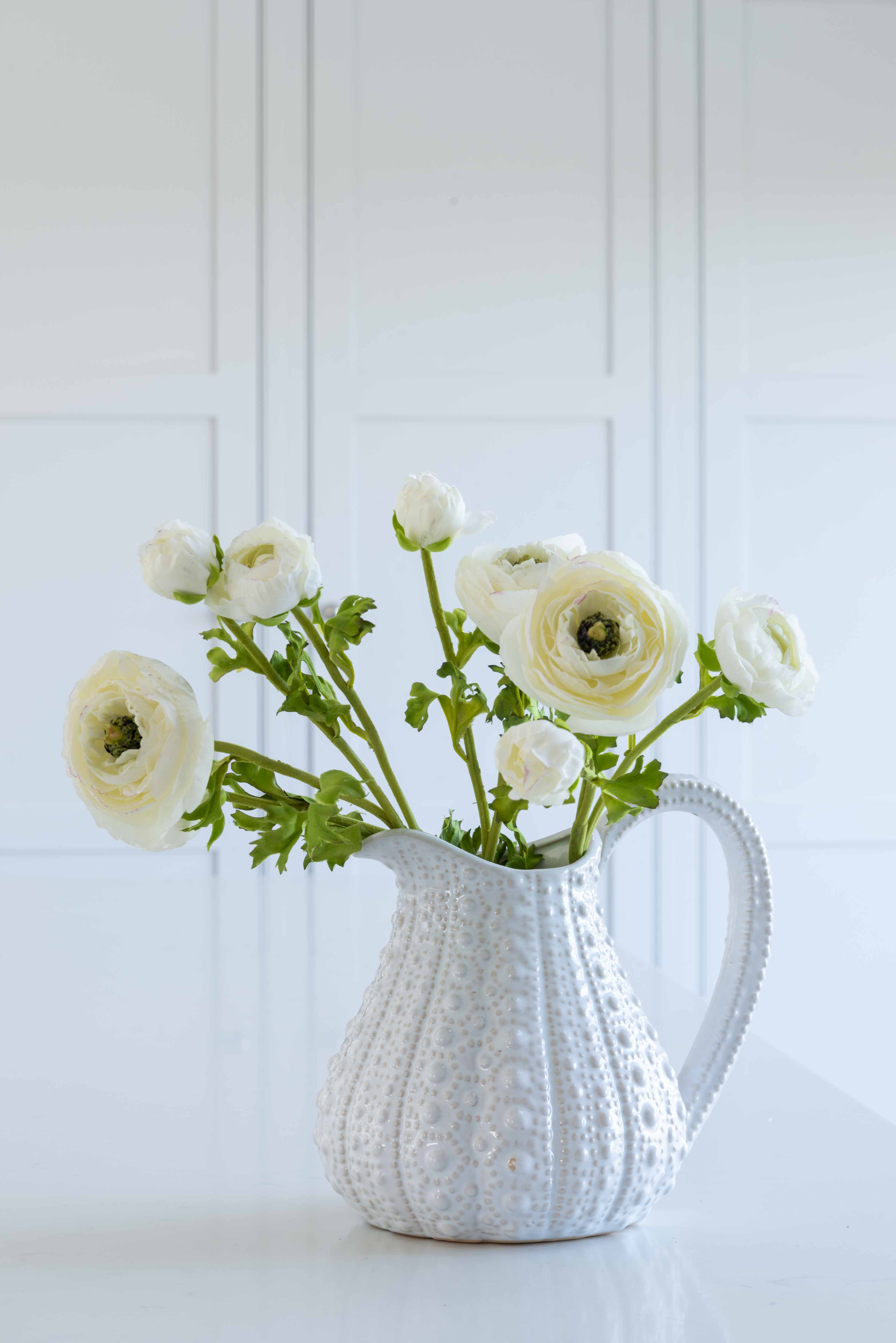 White ceramic pitcher holding realistic faux white ranunculus flowers with green leaves