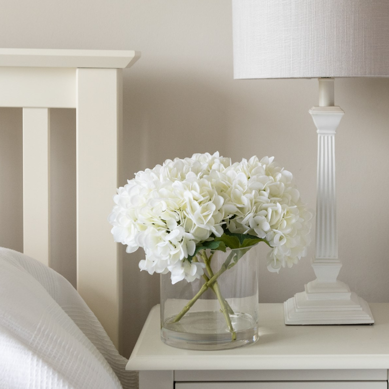 Faux white hydrangea arrangement in a clear glass vase with realistic stems and foliage