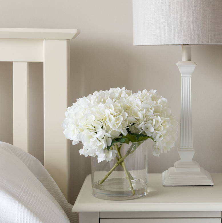 Faux white hydrangea arrangement in a clear glass vase with realistic stems and foliage