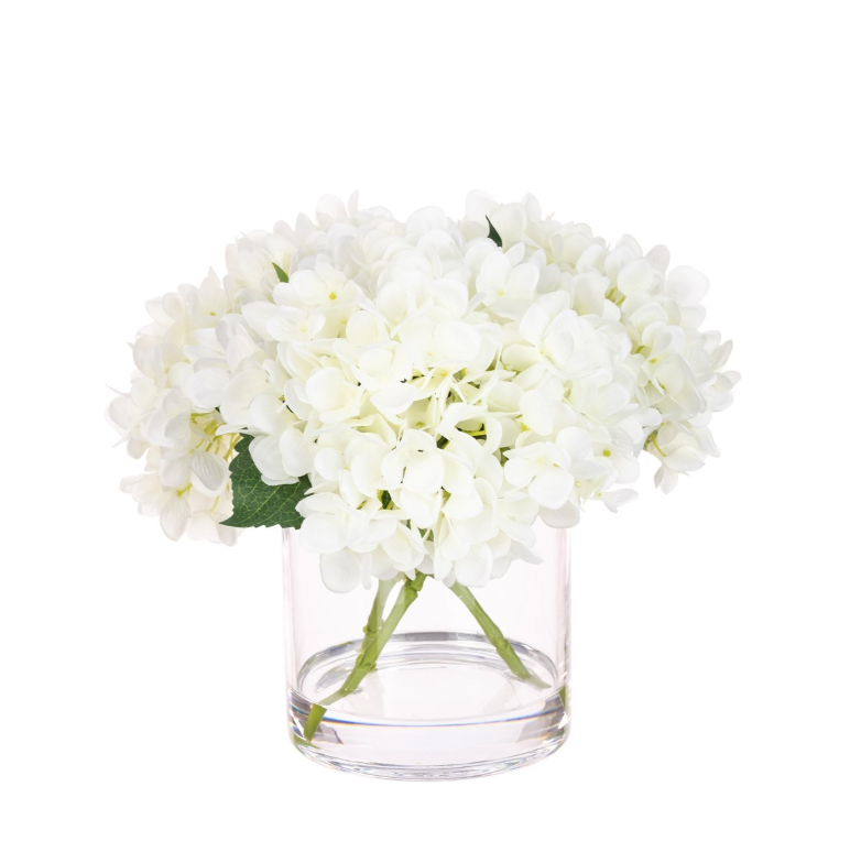 Faux white hydrangea arrangement in a clear glass vase with realistic stems and foliage