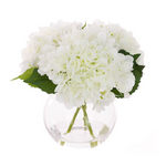Faux white hydrangea arrangement in a clear globe glass vase with realistic stems and foliage