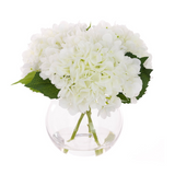 Faux white hydrangea arrangement in a clear globe glass vase with realistic stems and foliage