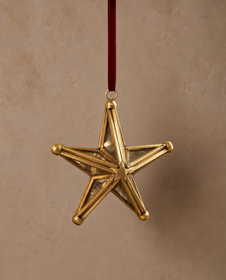 A five-pointed star Christmas ornament with a polished gold finish and a dark red hanging ribbon, shown against a textured beige background.