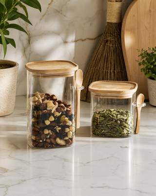 Two clear glass canisters with light wood lids and small wooden spoons, filled with mixed nuts and pumpkin seeds, styled on a marble countertop with kitchen decor in the background.