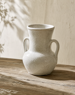 A handmade ceramic vase with two handles, featuring a textured off-white glaze, displayed on a rustic wooden table against a textured beige wall with soft leaf shadows.