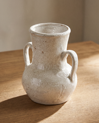A handmade ceramic vase with a textured, speckled off-white glaze and two loop handles, sitting on a wooden tabletop with warm natural light.