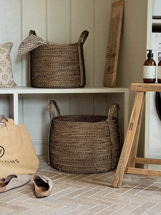 Two woven brown storage baskets with handles are displayed in a styled home setting. One basket is placed on a white shelf next to a patterned pillow, and the other sits on the floor.