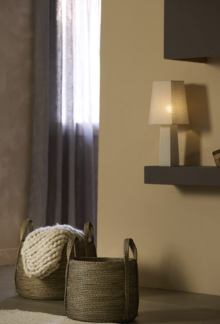 Two woven natural fiber storage baskets with handles, one holding a chunky knit cream throw blanket, set in a warm, modern room next to a window with gray curtains and a lit table lamp.