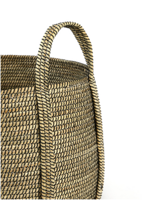 Close-up of a woven straw basket bag with a natural tan and dark brown interlaced pattern, featuring a single loop handle on a plain white background.