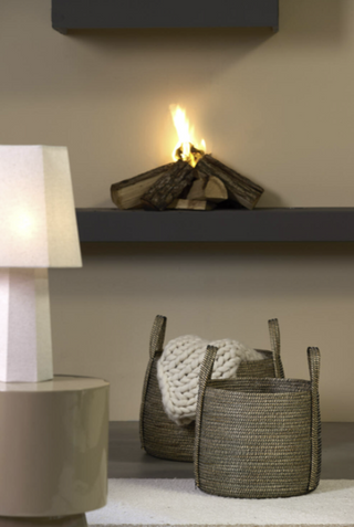 A styled interior scene with a lit fire burning in a decorative fireplace or shelf unit, alongside woven storage baskets, a white knit throw, and a lit table lamp against a warm beige wall.