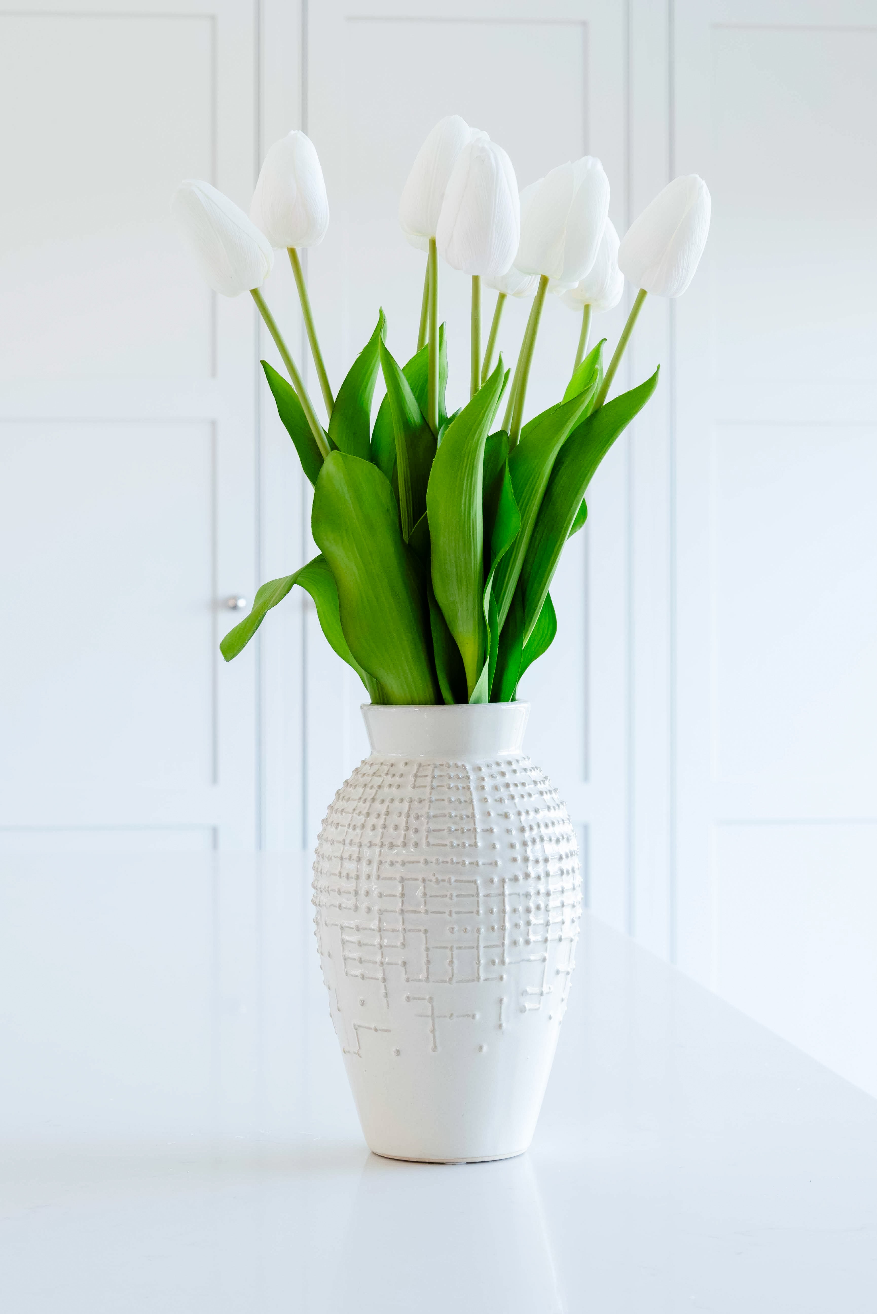 A stunning white ceramic vase holding white lilies. There is a raised-bump pattern on the exterior of the vase