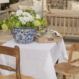 Outdoor setting with a table set for a meal, featuring a blue and white floral pot, white flowers, and wooden chairs.