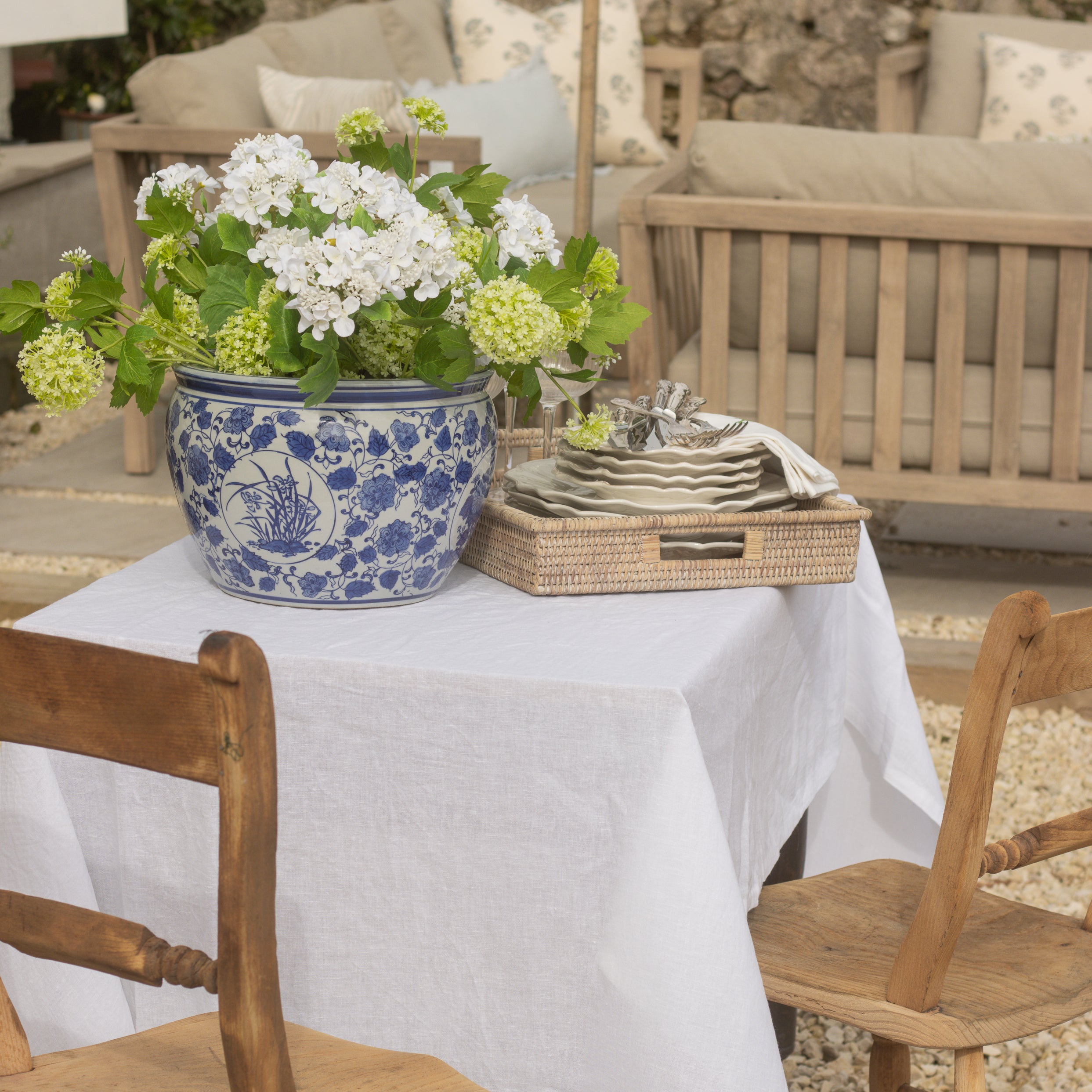 Outdoor setting with a table set for a meal, featuring a blue and white floral pot, white flowers, and wooden chairs.